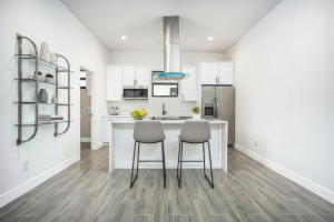 Modern white kitchen with island, two gray bar stools, stainless steel appliances, and glass shelving.