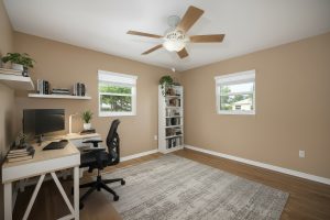 Home office with beige walls, wood floor, white desk, black chair, bookshelf, and ceiling fan.