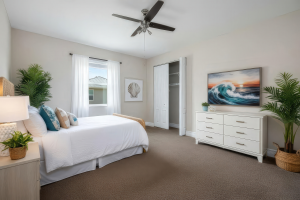 Bedroom with white bed, decorative pillows, window with curtains, open closet, white dresser, ocean wave painting, ceiling fan, and potted plants.