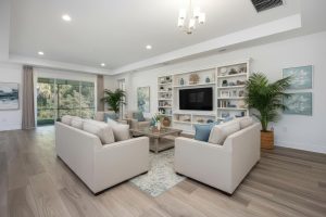 Light-toned living room with sofas around a coffee table, white shelving, TV, plants, and sliding glass doors.