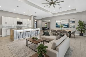 Open living space with modern kitchen, bar stools, dining table, sofa, coffee table, plants, and ceiling fan.